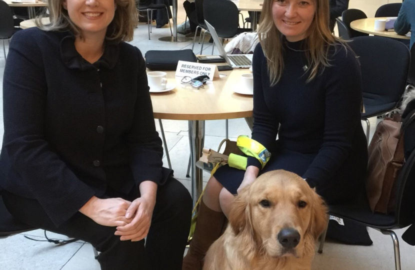 Lana Hempsall sitting in the atrium of Portcullis House with Penny Mordaunt MP and Zorin in his working hguide dog harness