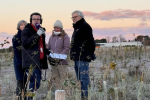 image shows news reporter with headphones and a microphone interviewing a group of people in a field covered with frost 