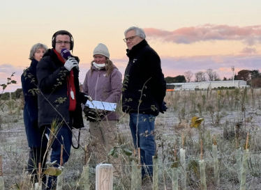 image shows news reporter with headphones and a microphone interviewing a group of people in a field covered with frost 